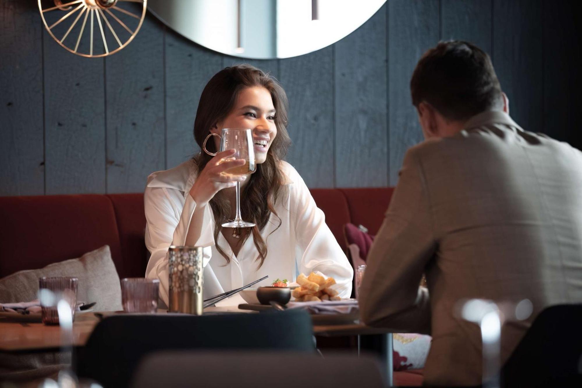 丽笙布鲁酒店天空酒店 塔林 外观 照片 The photo depicts a woman and a man sitting at a table in a restaurant. The woman is smiling and holding up a large glass, possibly containing a drink, while engaging in conversation with the man, who is seated across from her. The setting has a mode